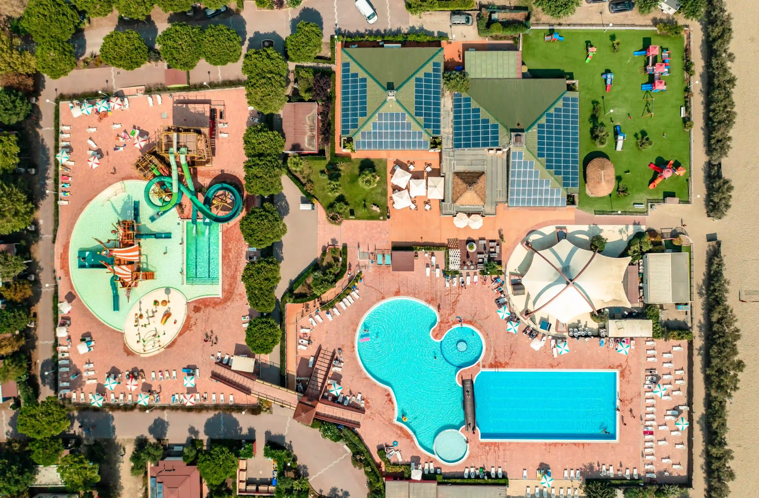 Veduta aerea di un complesso turistico con piscine, scivoli d'acqua, ombrelloni, lettini e aree verdi.