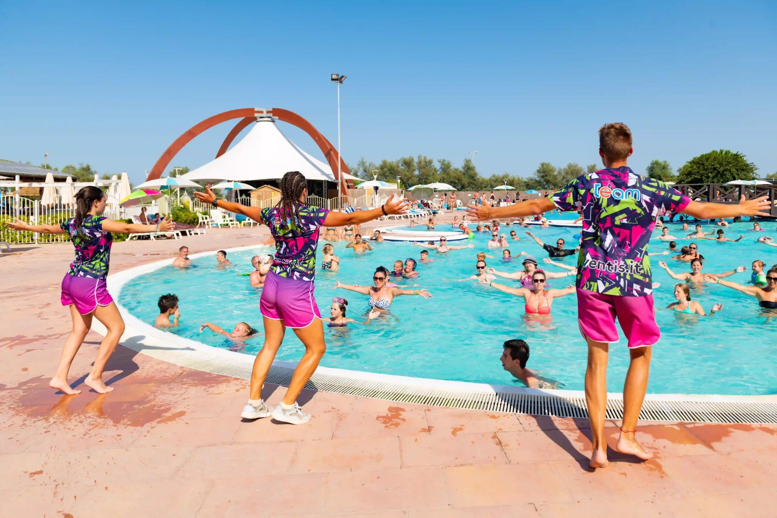 Animatori in abiti colorati che guidano una sessione di aerobica in acqua per una folla entusiasta in una piscina di un resort.